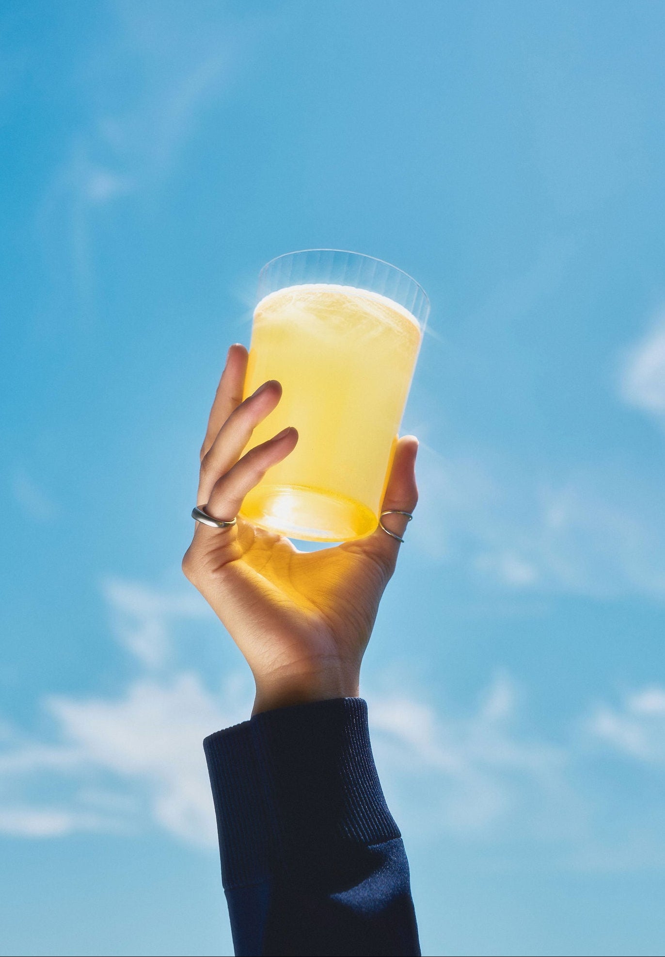 A hand wearing a dark navy sleeve holds a clear glass cup filled with a pale yellow beverage against a bright blue sky with light clouds.